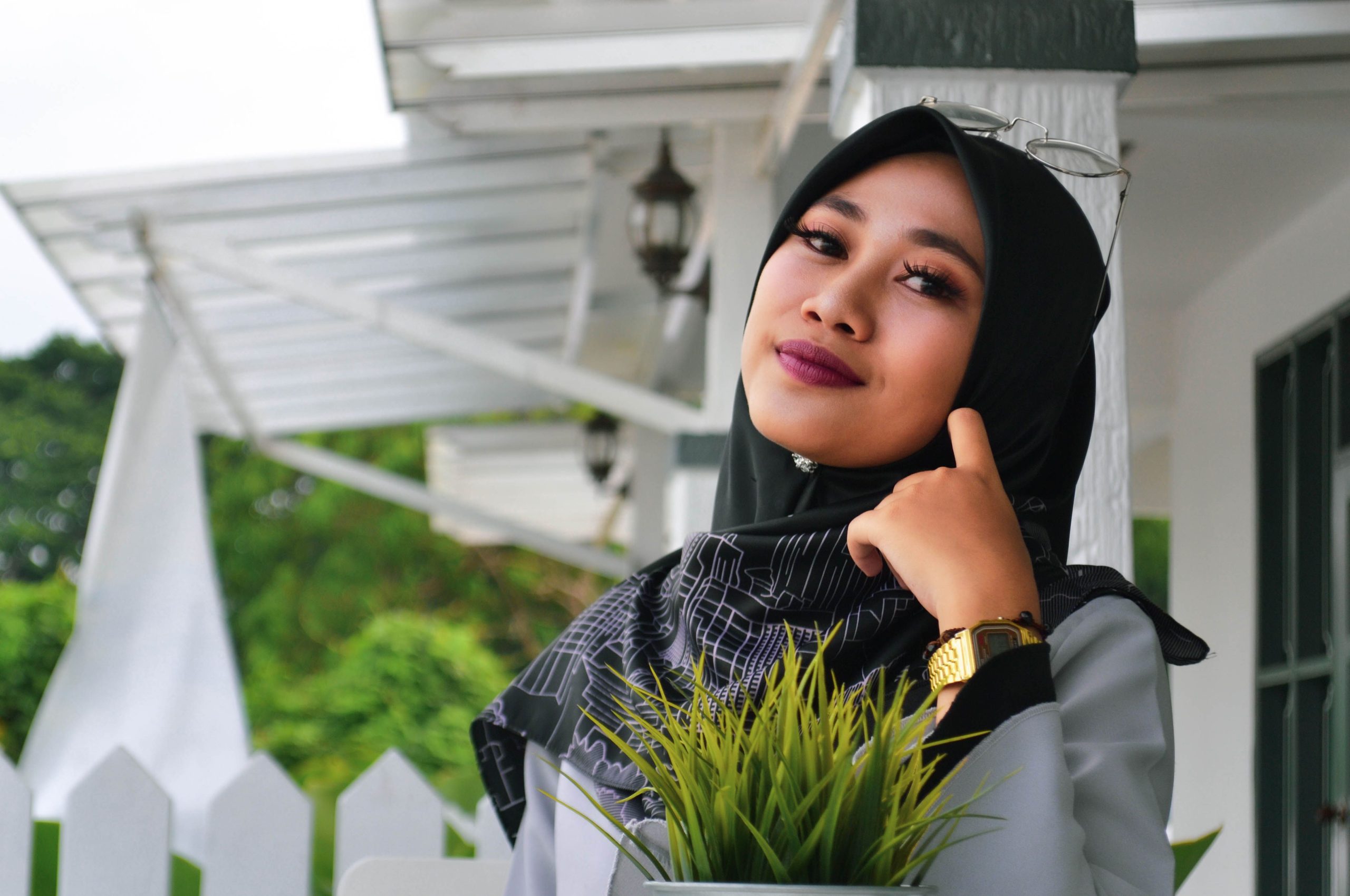 Elegant woman in hijab with makeup posing outdoors in front of a white house, exuding confidence and style.